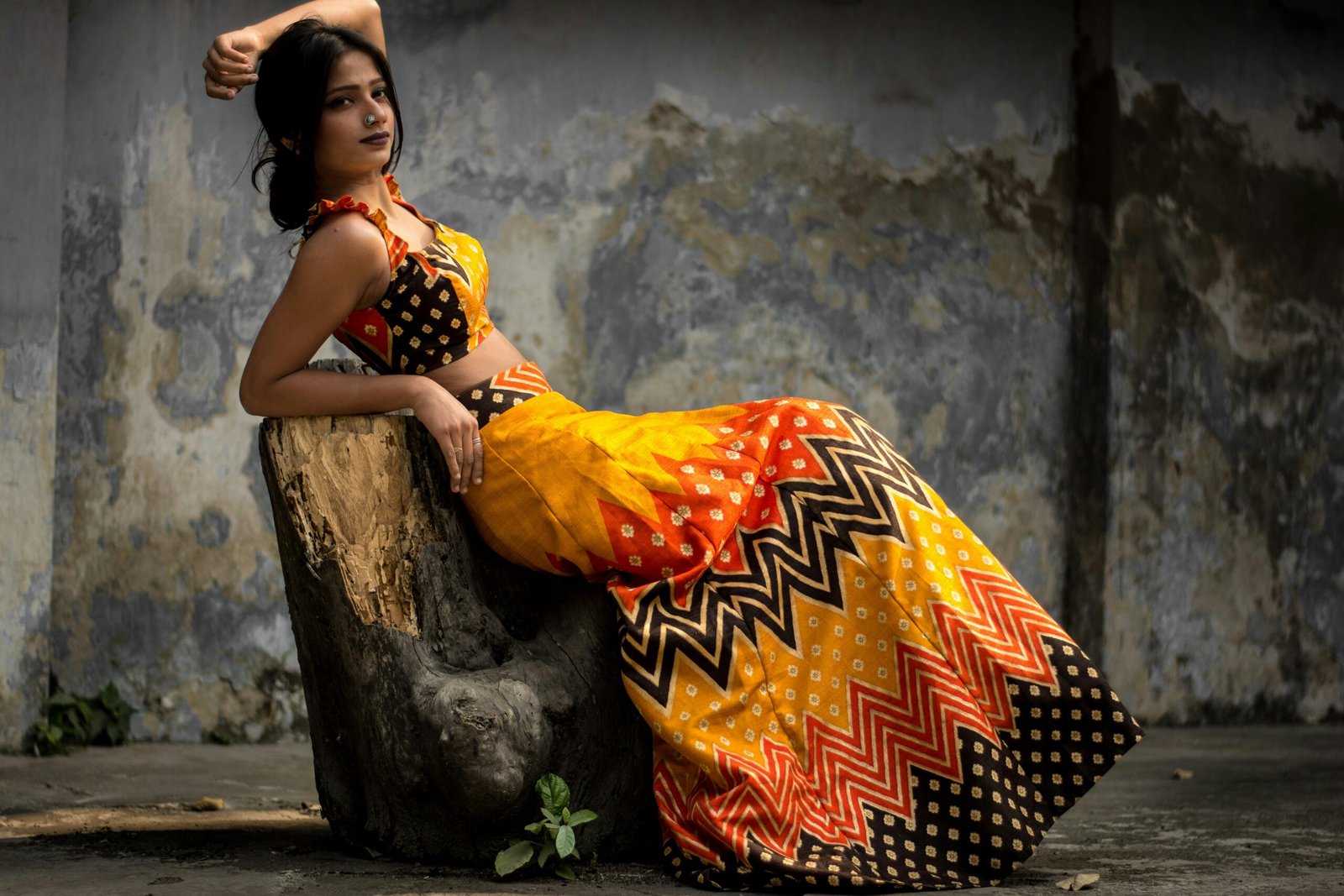A woman elegantly poses on a tree stump wearing a colorful traditional outfit.