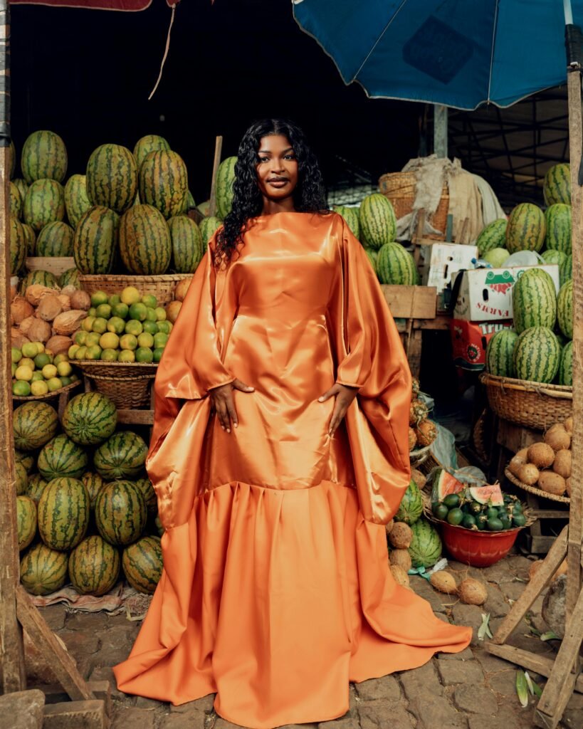 Fashionable woman in an orange gown poses in a colorful fruit market. Lively scene with melons and limes.
