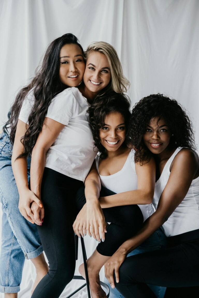 Four diverse women embrace, smiling, showcasing friendship and unity indoors.
